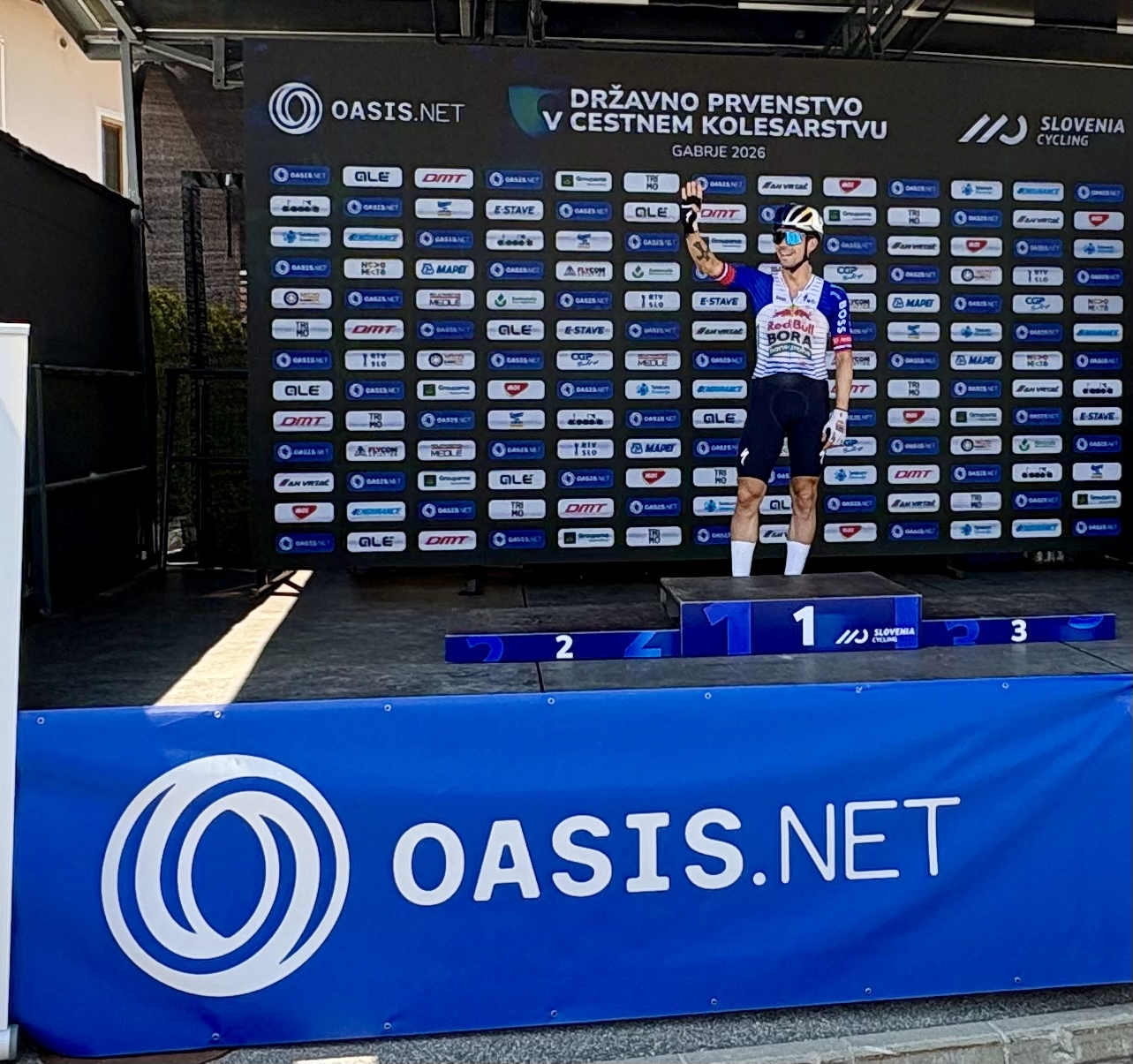 A rider waving from the winners' podium at the Slovenian Nationals, backed by the Oasis sponsor wall
