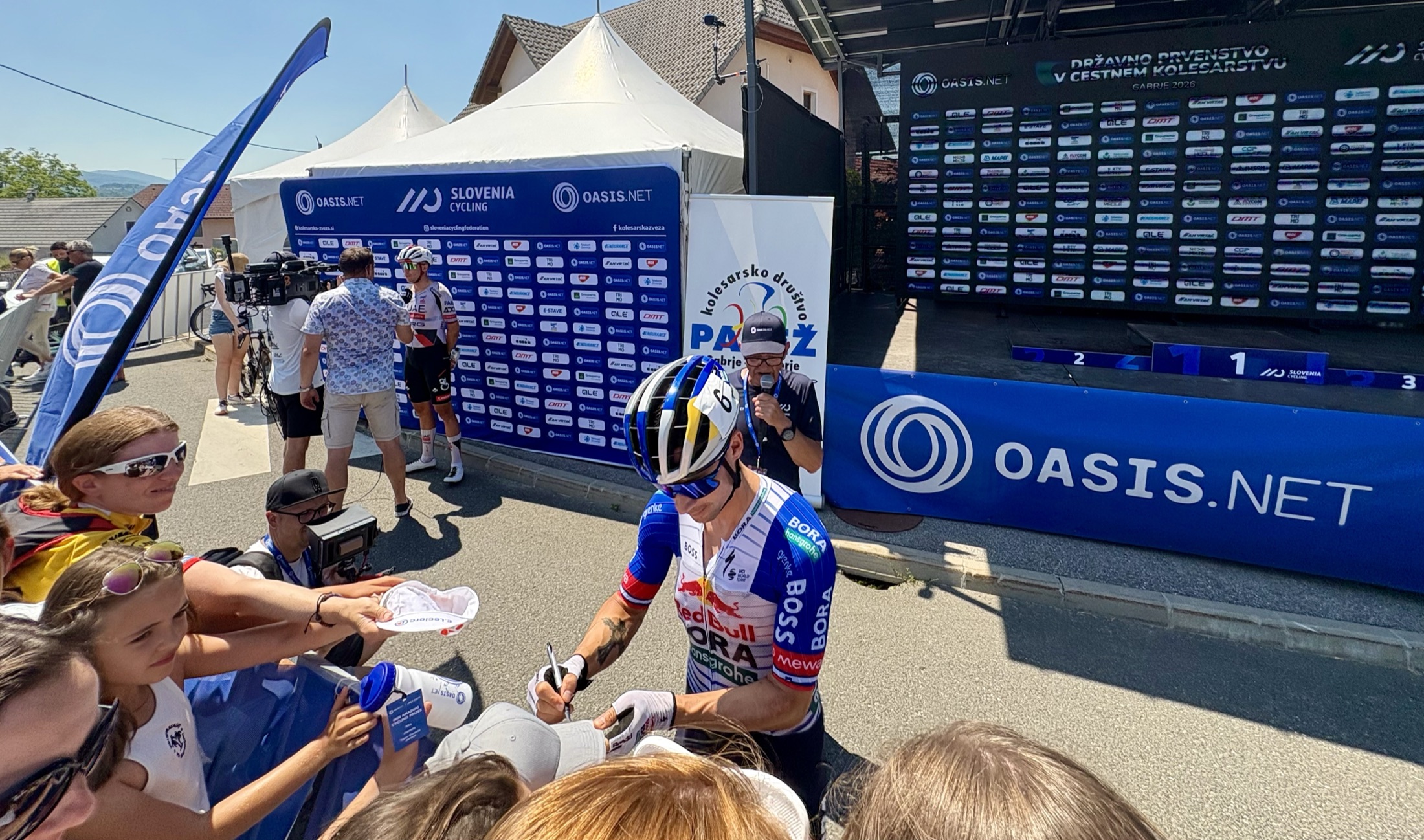 A rider signing autographs for young fans beside Oasis-branded barriers in Gabrje